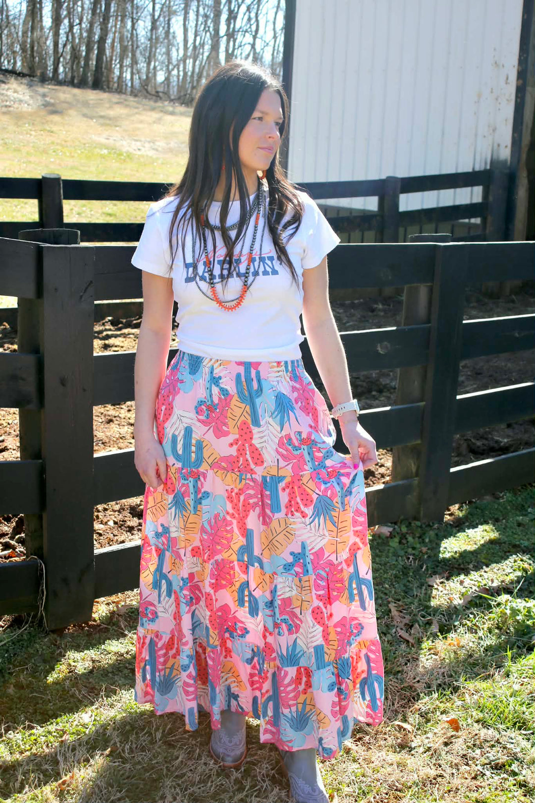a beautiful lady in a western skirt, western jewelry, and cowboy boots on a farm near a fence and barn.