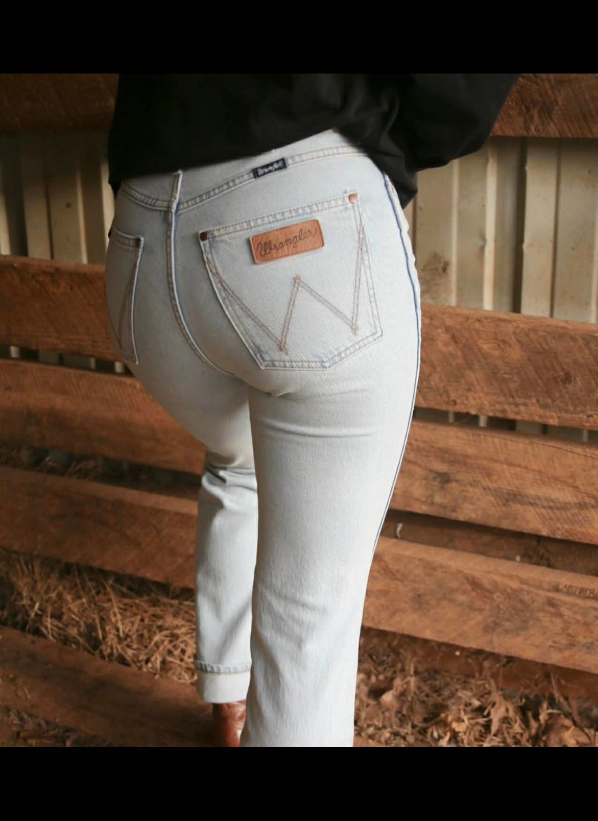 Person wearing light blue jeans with a brand logo in a barn setting wrangler