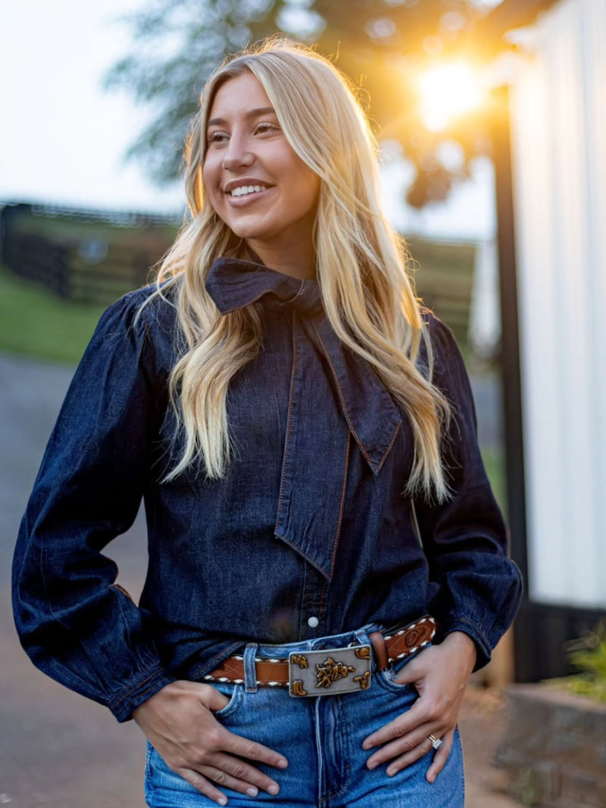 A smiling blonde woman wearing a dark navy long-sleeve denim shirt with a large bow collar and blue jeans, standing outdoors in warm sunlight.